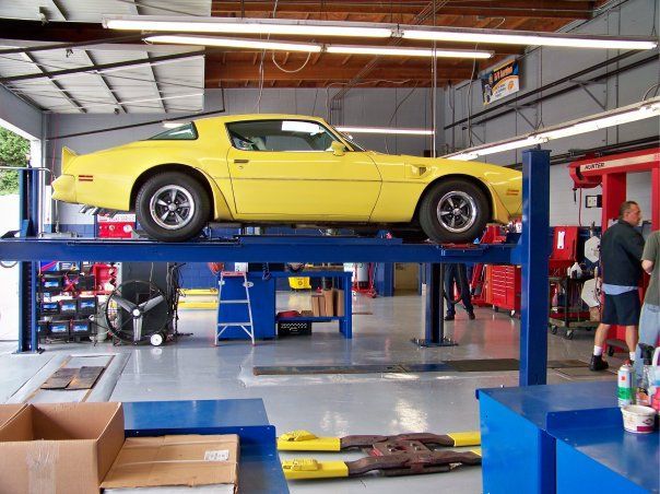 A bright yellow classic sports car lifted on a blue hydraulic shop rack inside a garage with two people working nearby.