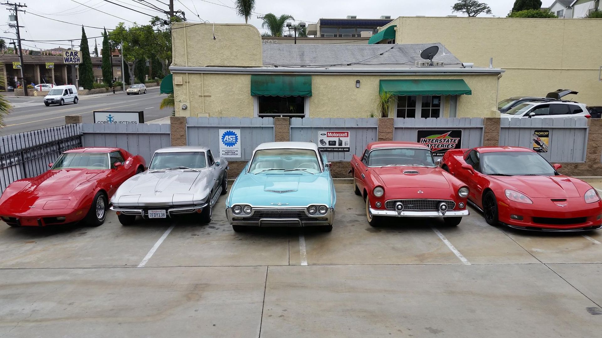 Five classic and modern sports cars, including red, silver, and light blue models, parked in a row outside a building.