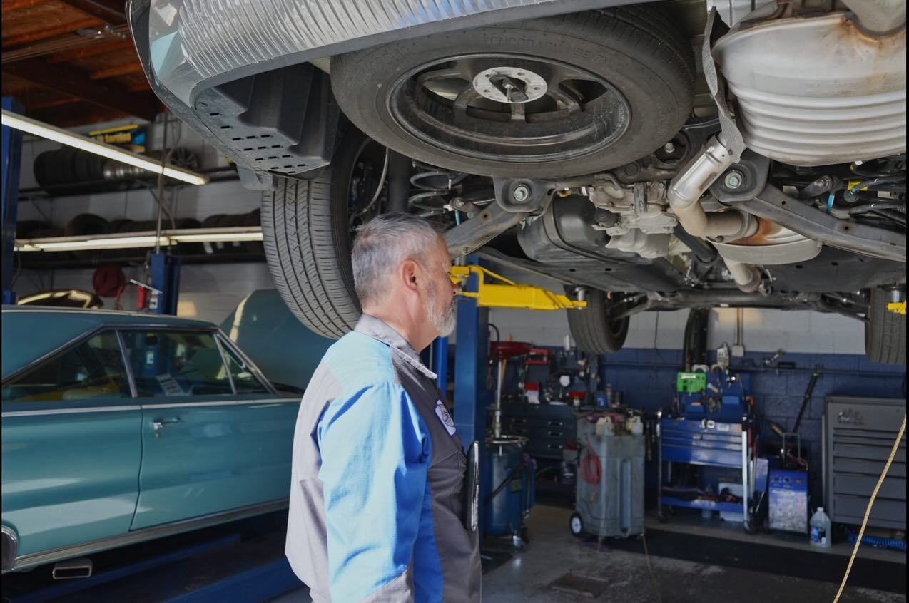 A technician inspects the undercarriage of a vehicle lifted on a hydraulic hoist inside an auto repair shop.
