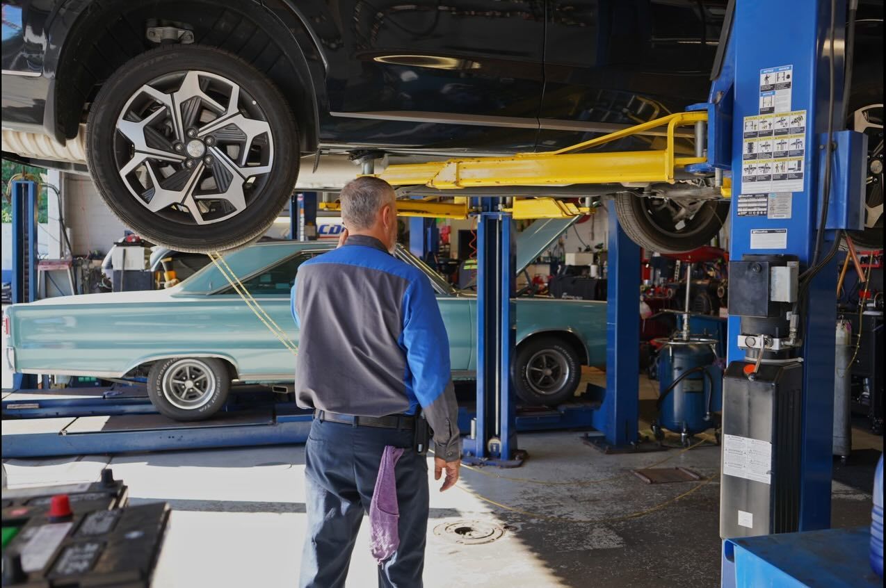 A technician stands in a repair shop looking at a classic light blue car parked beneath a lifted modern vehicle.
