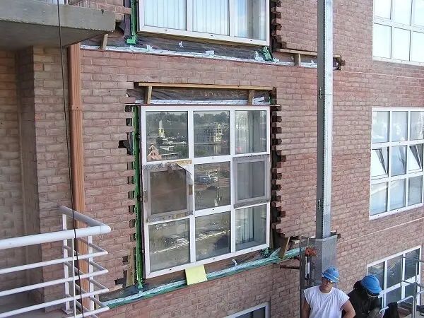 A man is standing in front of a brick building with a window being installed.
