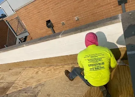 Person in neon yellow shirt and pink hoody on a roof, working. Brick building in background.