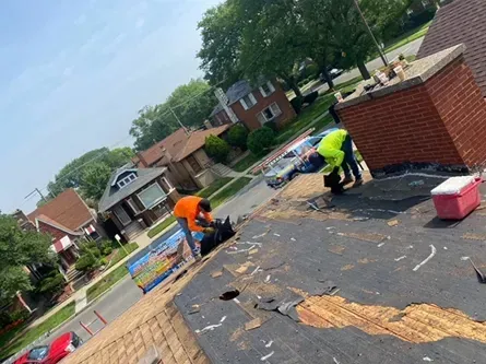 Roofers repairing a residential roof with visible damage, near a brick chimney and houses.