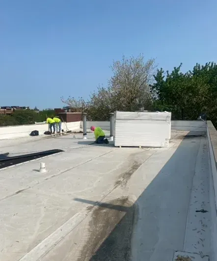Two workers in yellow vests on a white flat roof, near a vent and a small tree, working on roof repairs.