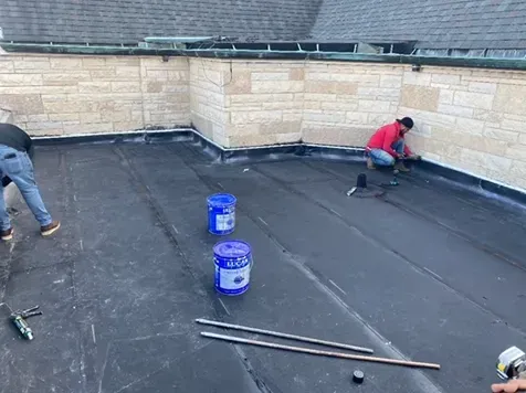Workers installing roofing on a flat, black roof near a brick wall. Blue buckets and tools are present.