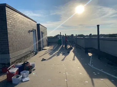 Rooftop scene with workers near brick walls in bright sunlight. Coolers, tools scattered on a flat surface.