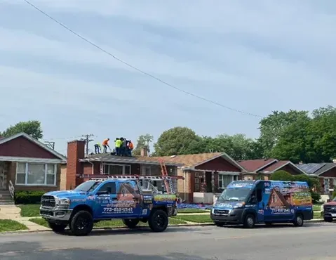 Roofers working on a house; blue truck and van with company logo parked on the street.