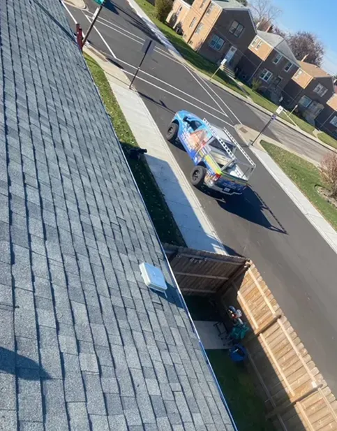 Blue truck on a street next to a house with a gray shingled roof, on a sunny day.