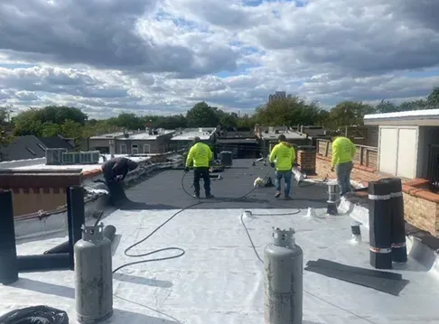 Roofers in yellow vests on a flat roof, installing roofing material. Propane tanks and a cloudy sky visible.