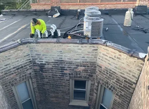 Roofer in yellow vest working on a black tar roof next to a brick wall with windows.