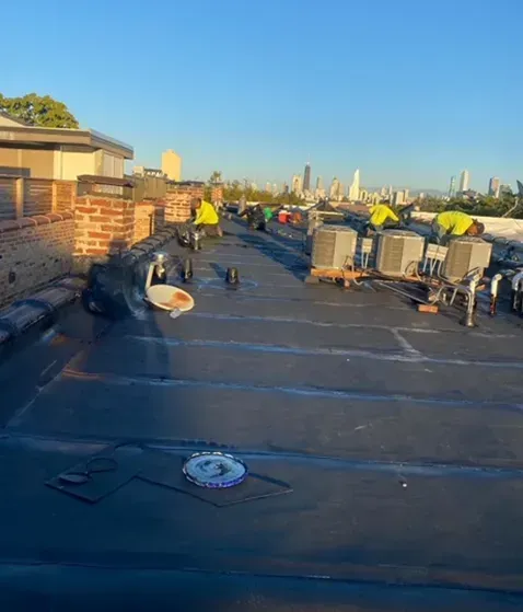 Workers in yellow vests on a black flat roof with HVAC units, cityscape background.