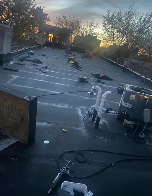 Flat rooftop with tools, equipment, and plumbing vents scattered; sunset in the background.