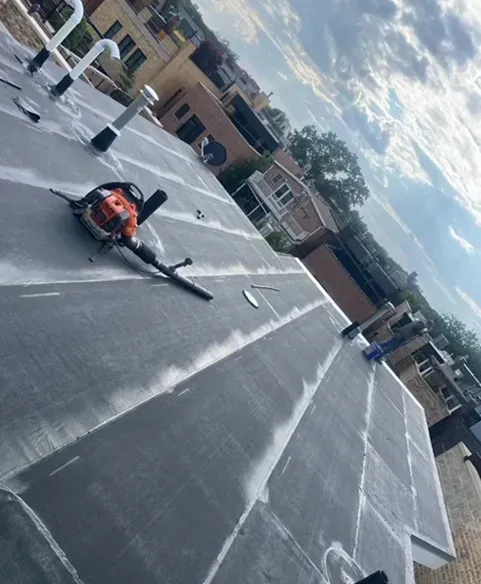 Rooftop with black tar sections; an orange blower lies on the surface. Gray sky and buildings in the background.
