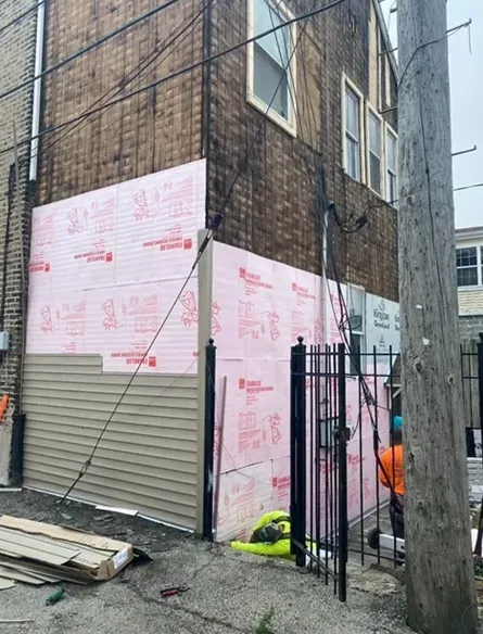 Building exterior under construction; pink insulation, tan siding. Worker near black gate.
