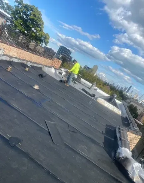 Roofer in safety vest working on a black, flat roof. Skyline and blue sky with clouds in the background.