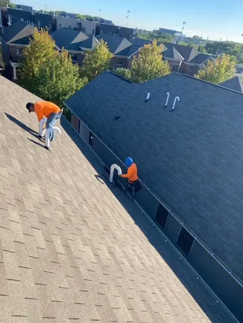 Two roofers working on a rooftop, one with an orange shirt and one with a blue hat.