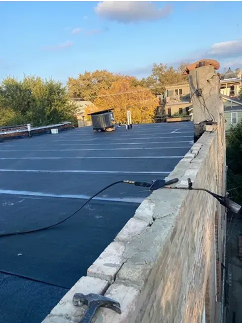 Flat, black roofing with a brick wall and chimney, a hammer, and trees in the background under a blue sky.