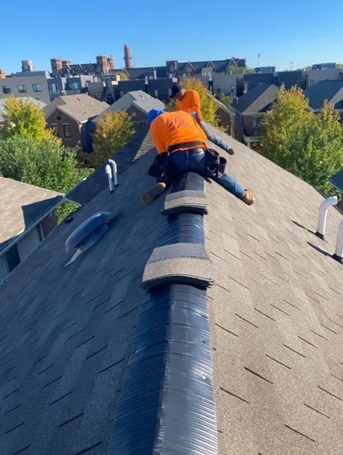 Two roofers in orange shirts working on a rooftop with a sunny, clear sky in the background.
