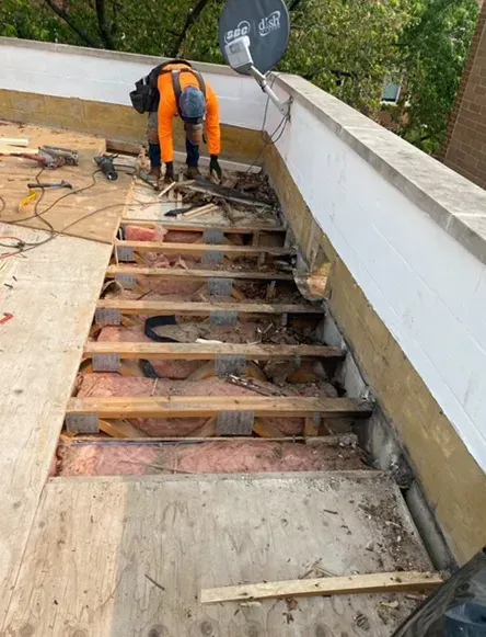 Person in orange workwear on a flat roof, removing old decking. Rotted wood and insulation visible.