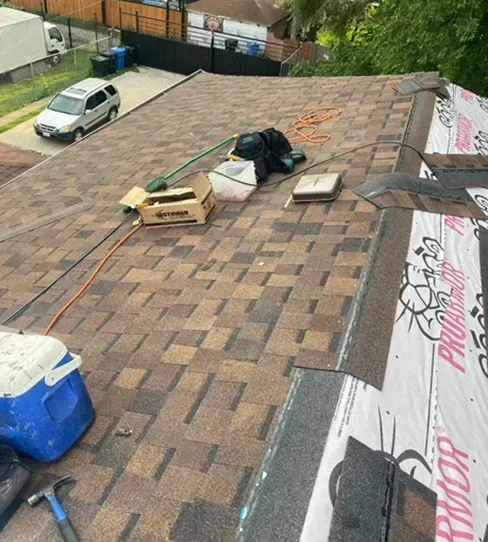 Rooftop with brown shingles, tools, and a blue cooler. Shingles partially installed, showing underlayment.