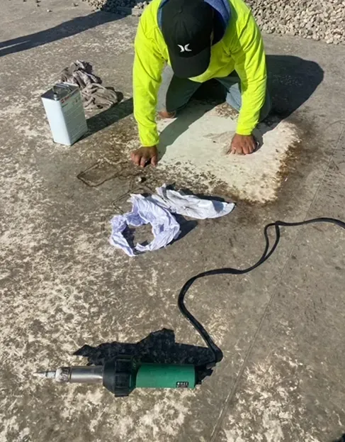 Person kneels repairing a roof. He wears a yellow shirt and black hat. A heat gun and solvent are nearby.