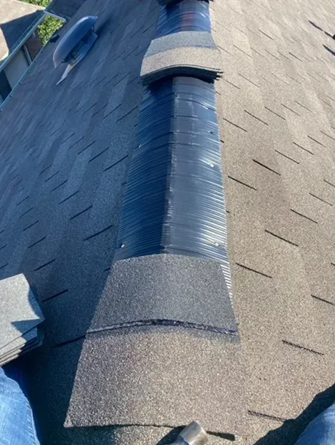 Roof with ridge cap being repaired, showing layers of black sealant and shingles.