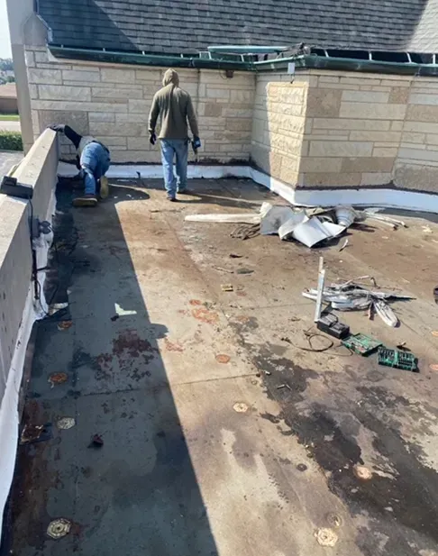 Two people working on a flat roof next to a stone building. Debris and tools are scattered.
