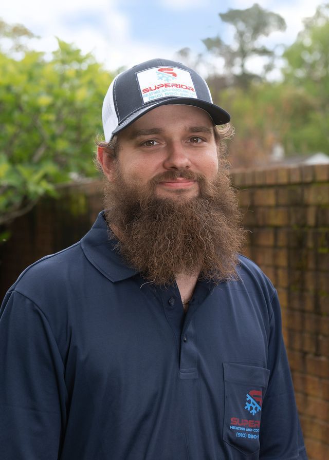 Man in work uniform and cap, smiling. Leaning against a brick wall, with greenery in the background.