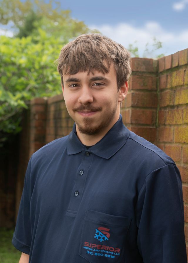 Man in a blue polo shirt smiles, stands near a brick wall and foliage. Shirt has a logo.