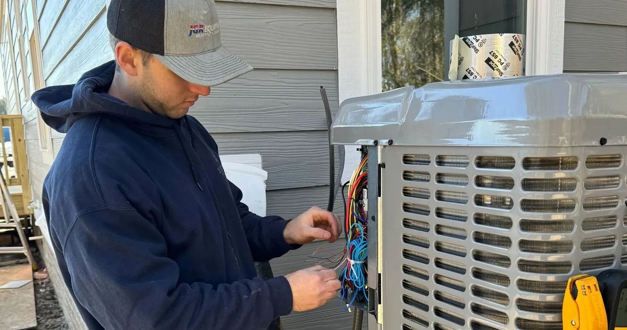 Man in a hat and hoodie wiring an air conditioner unit outdoors next to a house.