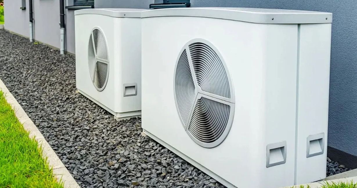 Two white heat pump units, gray gravel base, next to a building with green grass.