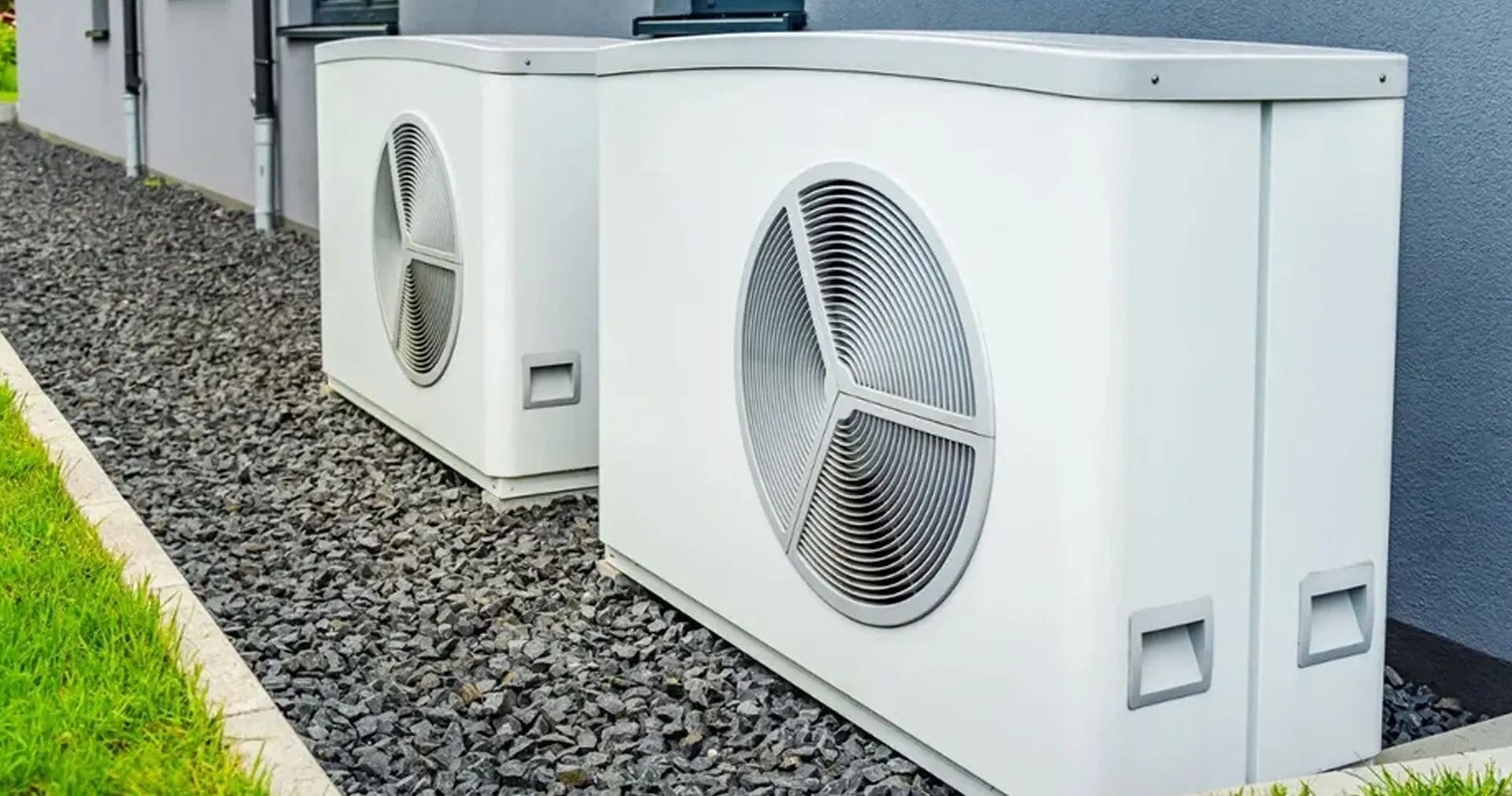 Two white heat pump units, gray gravel base, next to a building with green grass.