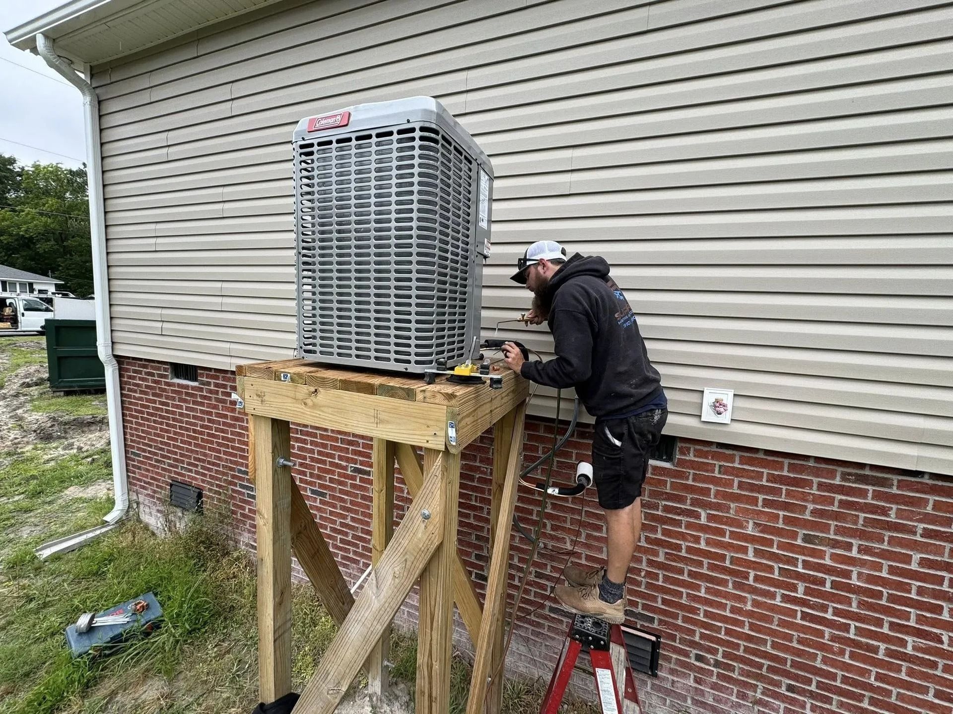 HVAC technician working on an air conditioning unit mounted on a wooden stand next to a brick house.