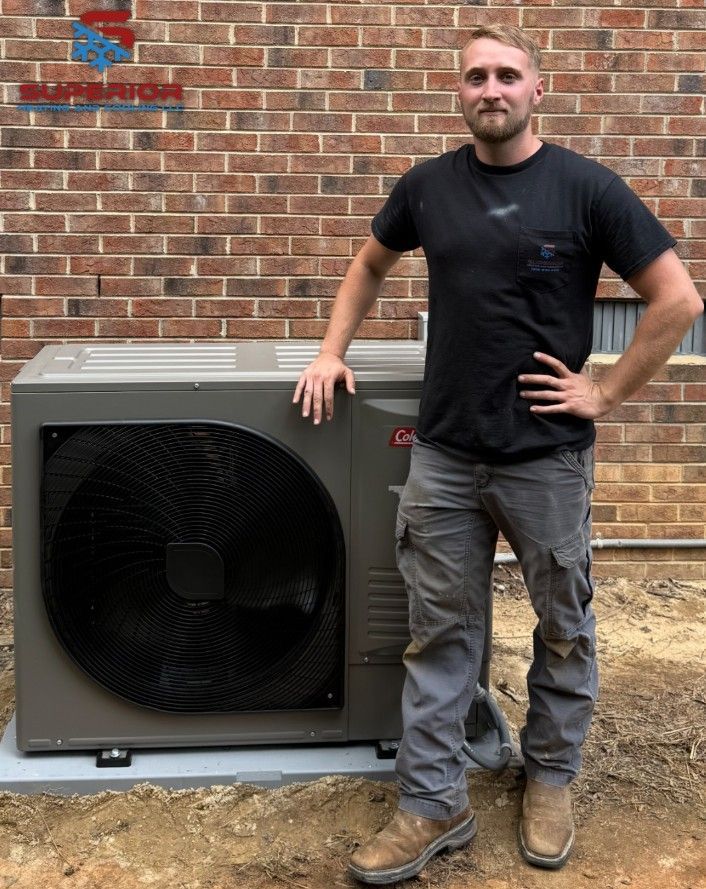 Man poses beside a new HVAC unit. He wears work clothes, stands in front of a brick wall and a concrete pad.