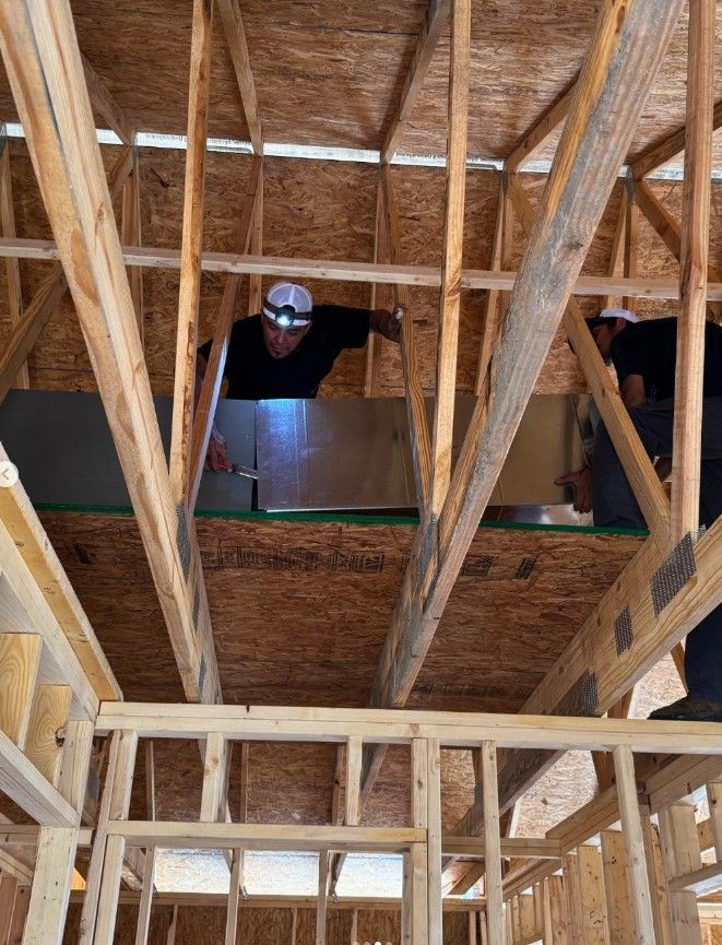 Two construction workers installing insulation in a wood-framed attic. One worker wears a headlamp.