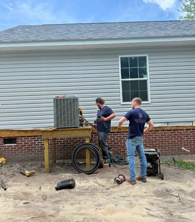 Two workers install an AC unit on a wooden platform against a gray-sided house.