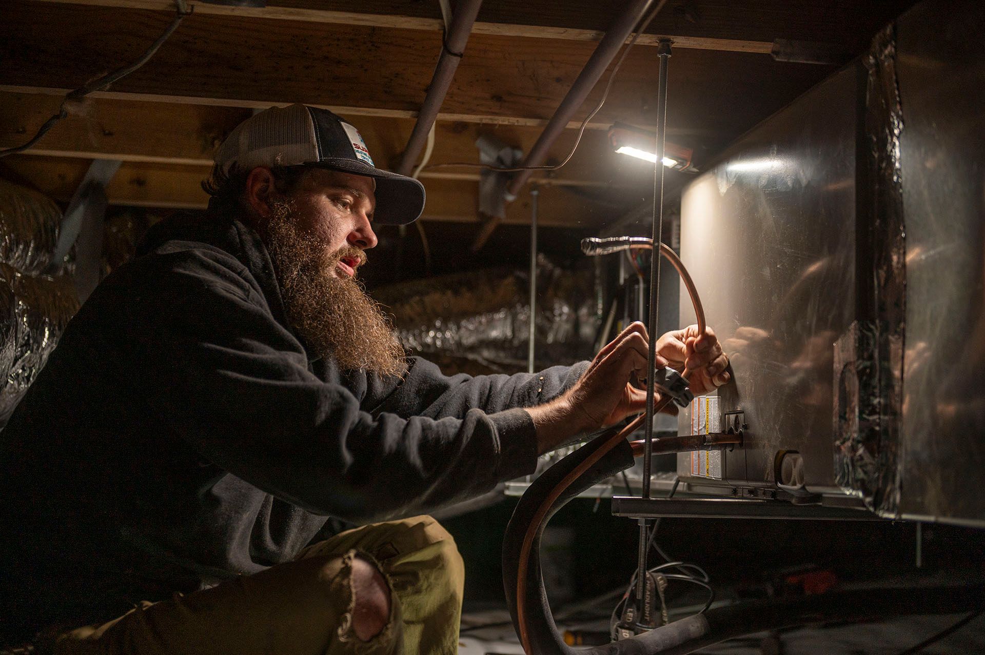 Man with a beard works on ductwork in a crawlspace, illuminated by a work light.