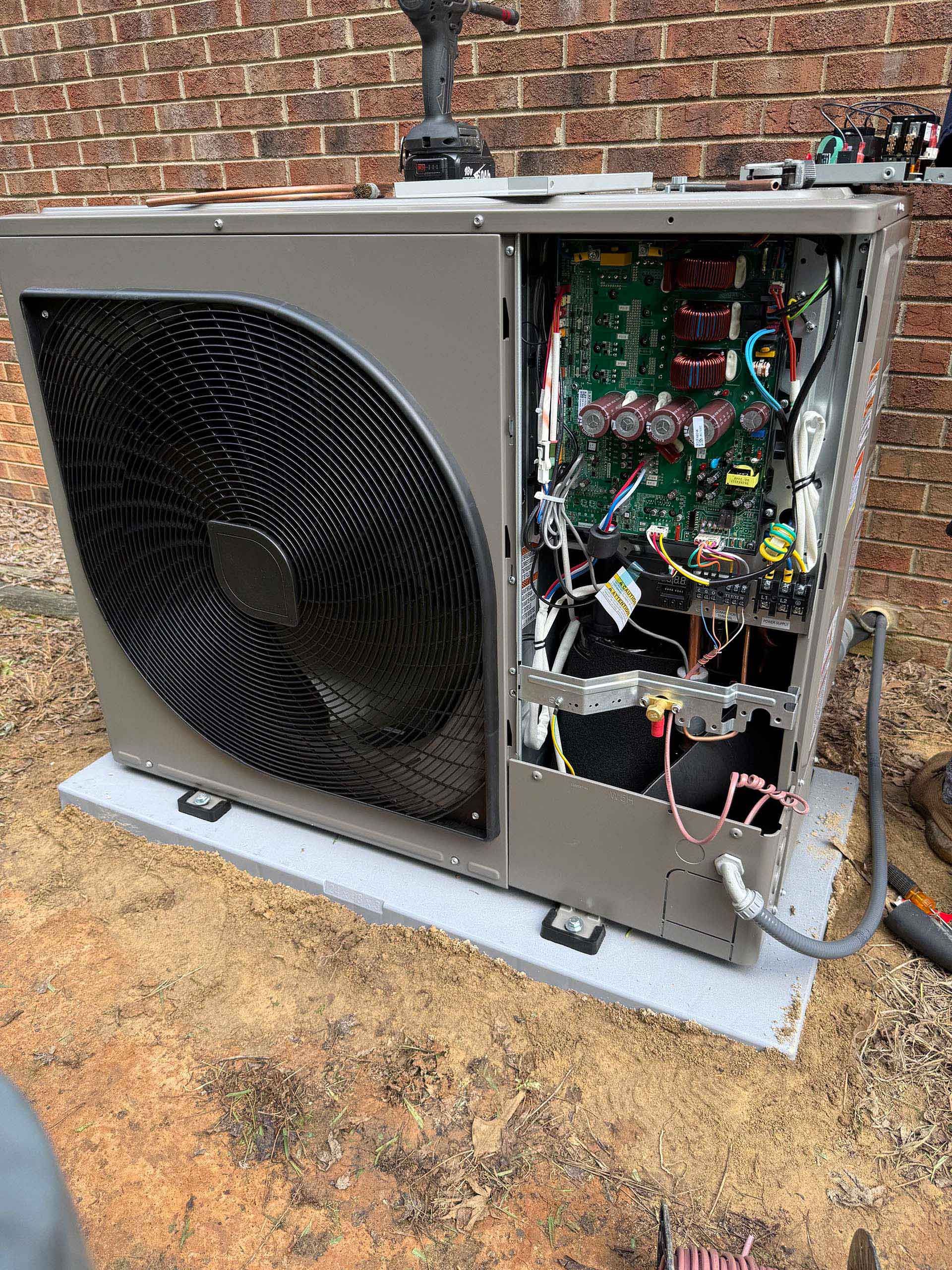 An air conditioning unit, partially disassembled, with circuit boards and wiring exposed, resting on a concrete pad.
