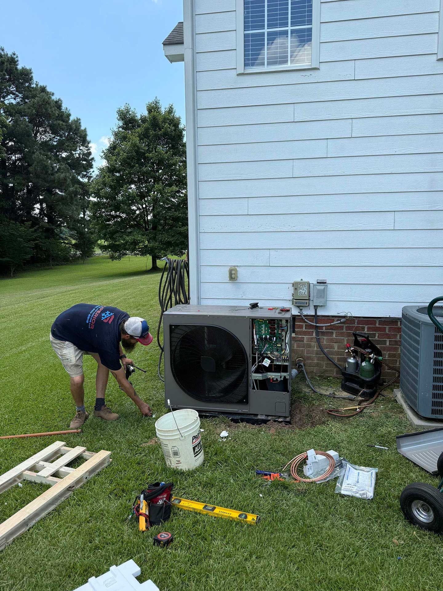 HVAC technician working on an AC unit outdoors.  The unit's panels are open. There is a house, tools, and yard nearby.