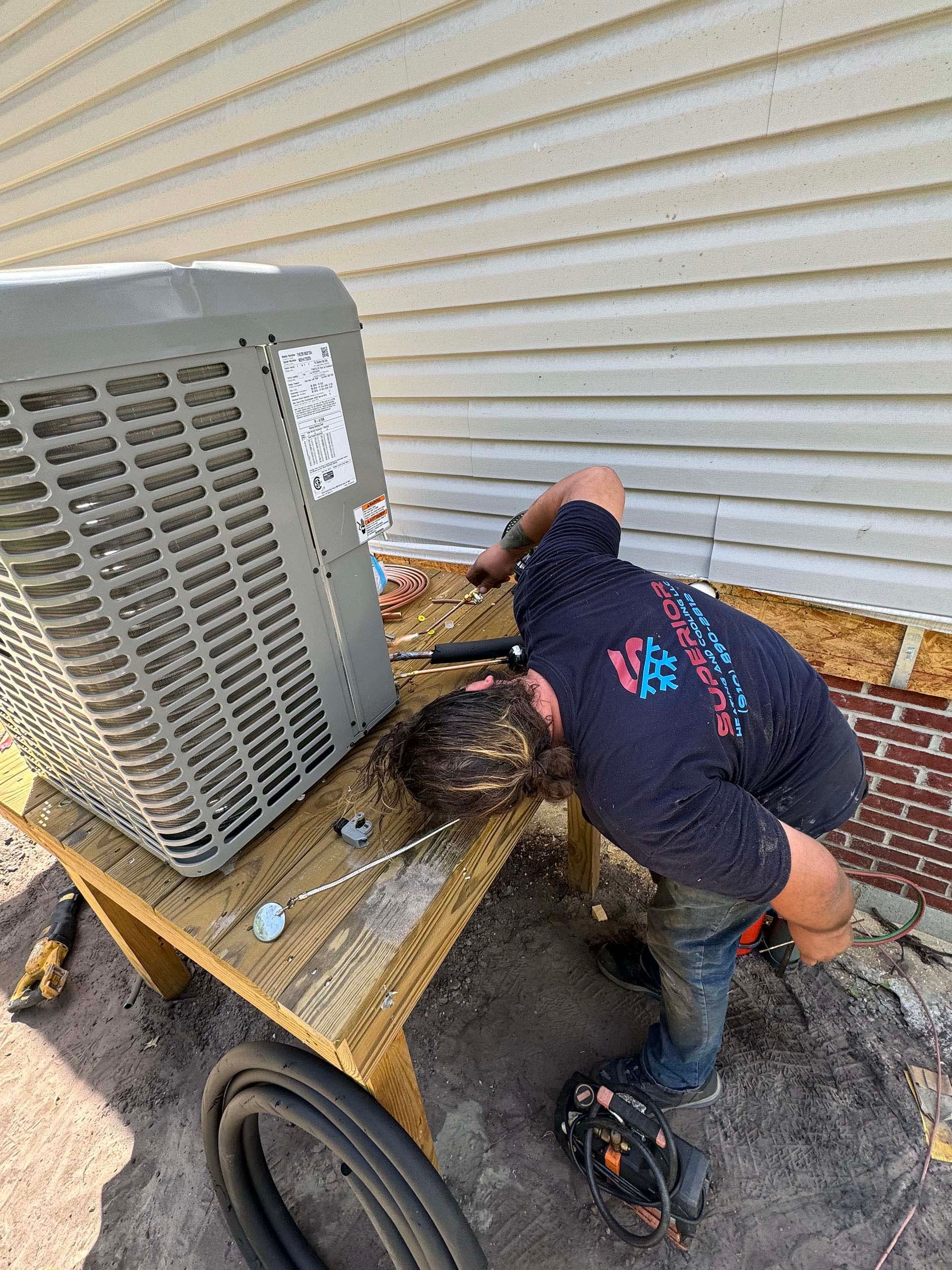 HVAC technician works on an air conditioning unit outside a building with beige siding.