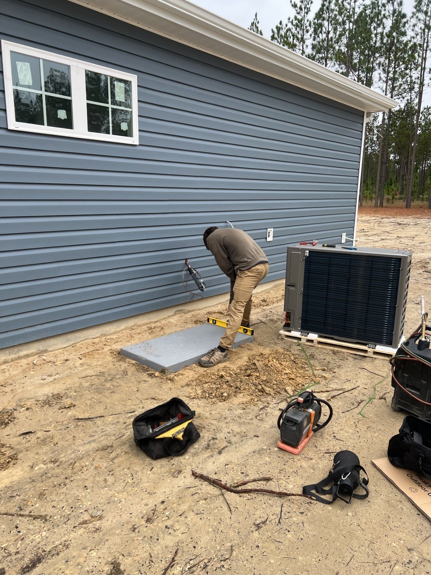 Person working on HVAC unit next to a blue house with window. Equipment and tools are scattered on the ground.
