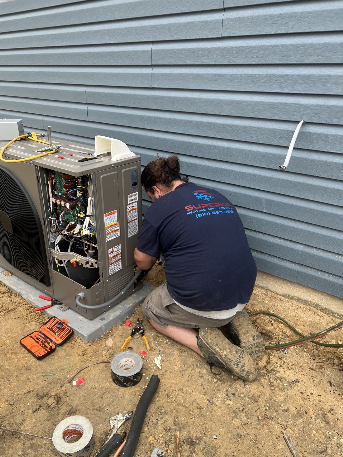 Person working on an AC unit outdoors. Gray unit, blue siding, tools on ground.
