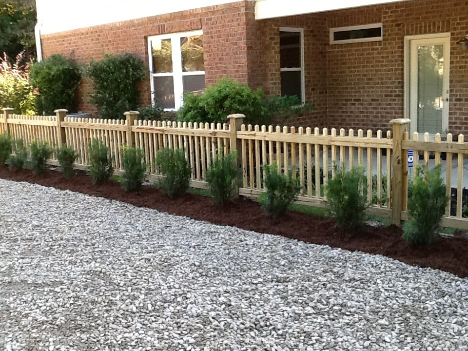 A wooden picket fence surrounds a gravel driveway in front of a brick house