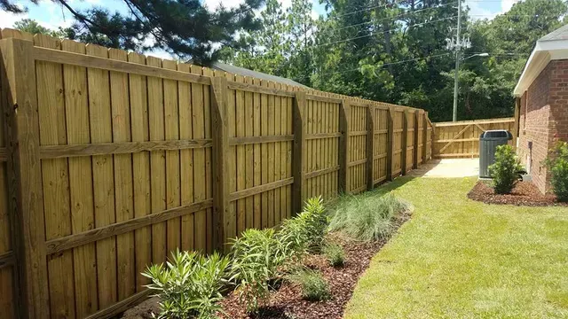 A long, light-brown wooden privacy fence runs alongside a grass lawn and a brick house wall with a small garden bed.