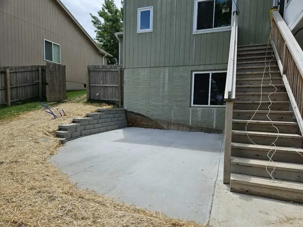 A newly poured gray concrete patio sits beside a green house with a wooden staircase and a small stone retaining wall.