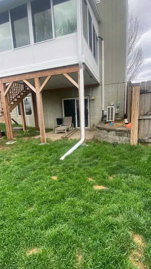 A view of a house backyard with an elevated porch supported by wooden beams, a sliding glass door, and a white downspout.