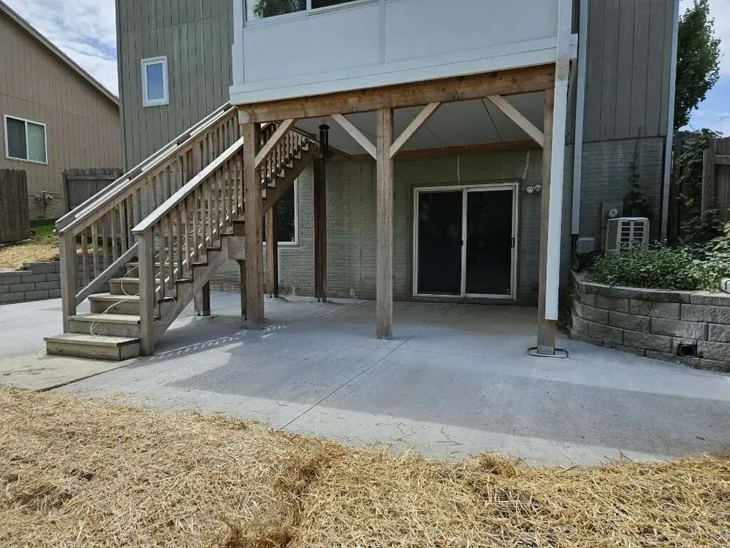 A concrete patio under a wooden deck with stairs leading to the upper level of a house.
