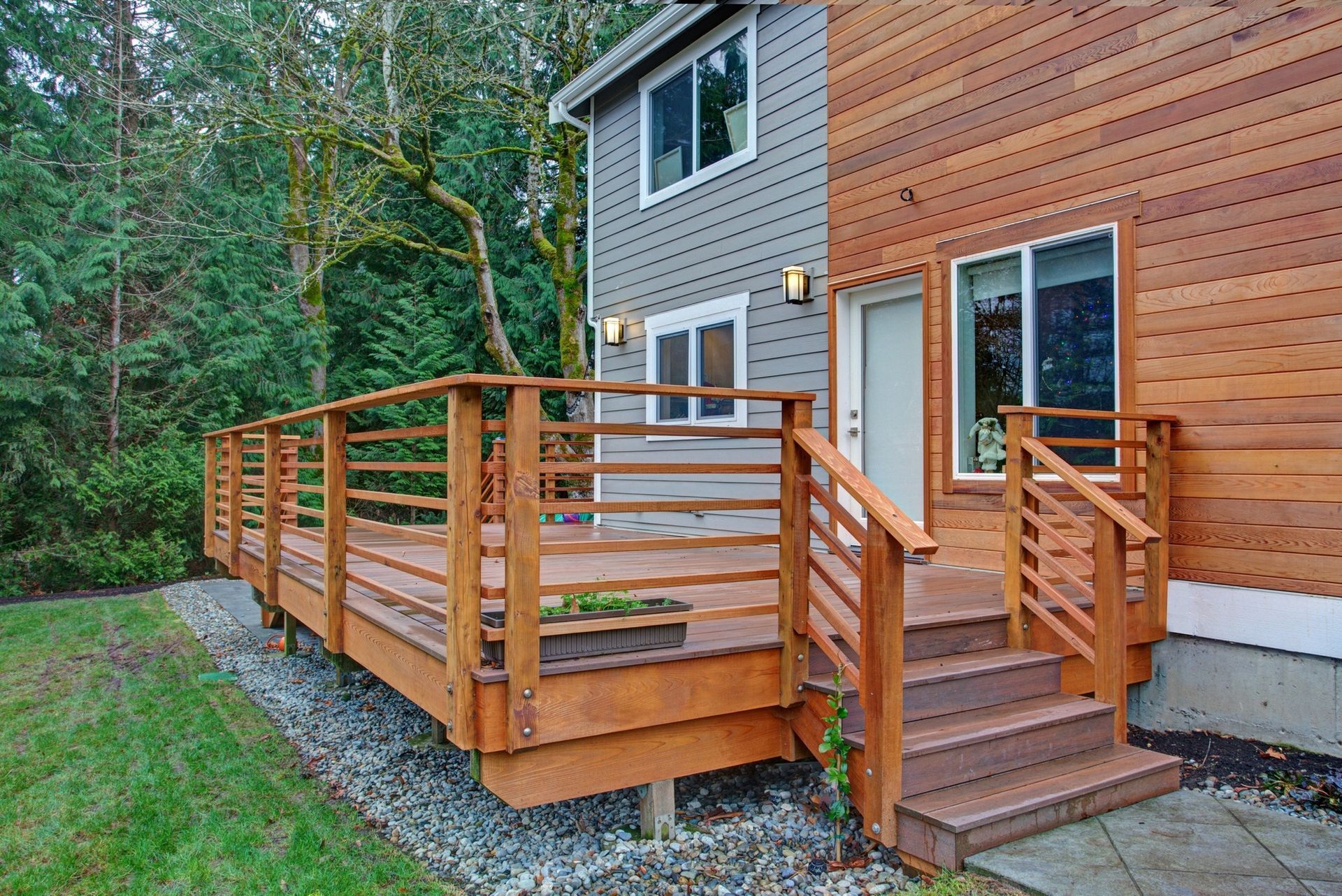 A wooden deck with horizontal railing attached to a two-story house with gray and wood siding, set against a forest edge.