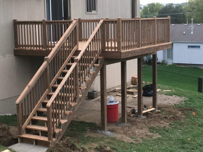A newly constructed wooden deck and staircase attached to the side of a tan house, overlooking a grassy backyard.