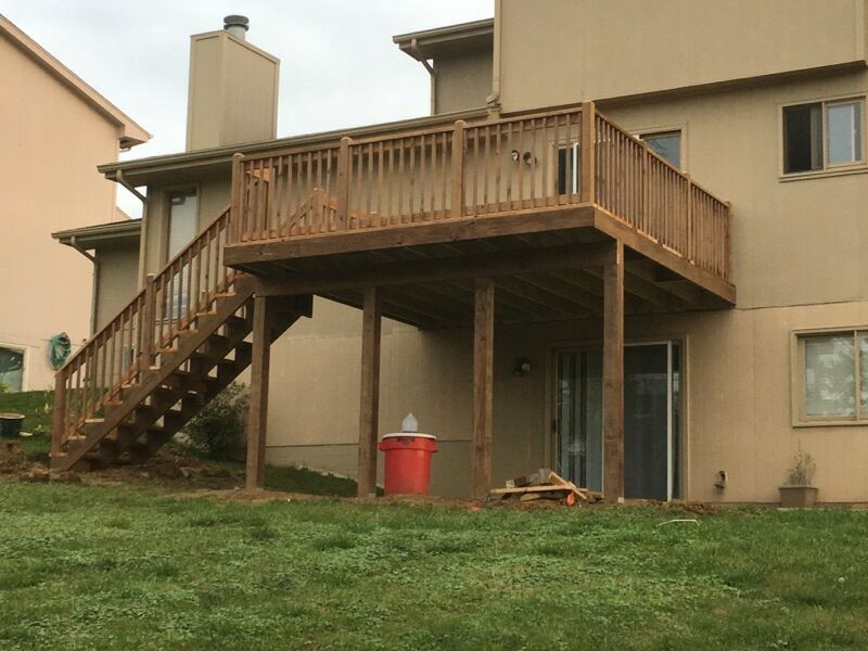 A tan two-story house with a wooden deck and staircase extending into a grassy backyard.